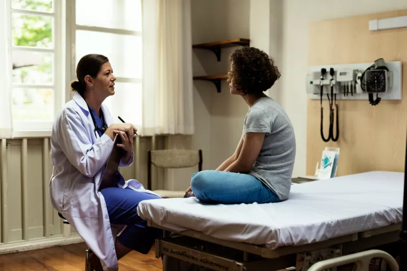 A young woman sits in a hospital room and talks about her health and wellness with her primary care doctor.