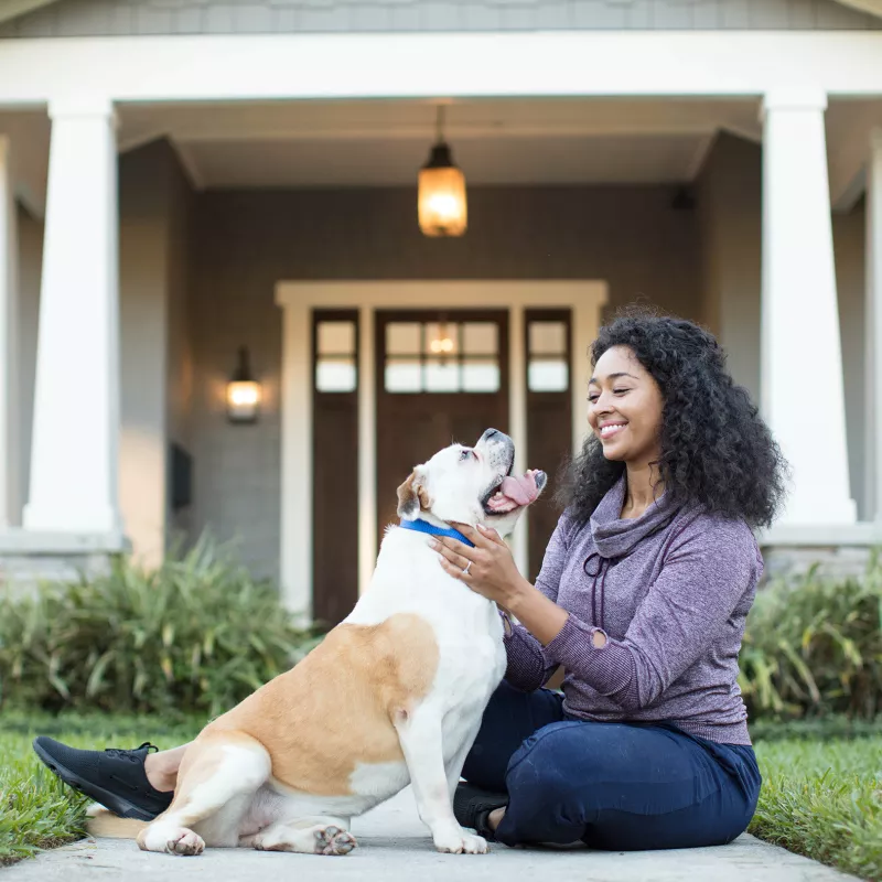 A young woman sits outside of a nice home, playing with her bulldog